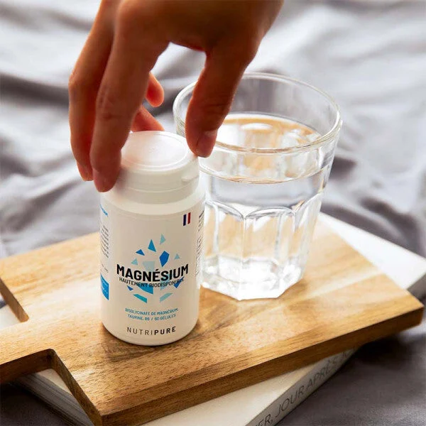 Une main saisit un flacon de magnésium Nutripure posé sur une planche en bois, à côté d'un verre d'eau